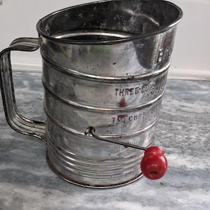 May include: Vintage silver-colored flour sifter with a red handle. The sifter has markings that read "Three Cups Measure", "Two Cups", and "One Cup".