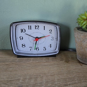 May include: A black and silver analog alarm clock with a white face, black numbers, and red and green hands. A small potted succulent plant sits to the right. The clock and plant are on a wooden surface.