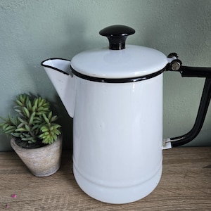May include: A white enamel coffee pot with a black handle, lid, and trim. The pot has a spout and a lid with a black knob. A small potted plant sits to the left of the coffee pot. The pot is on a wooden surface.