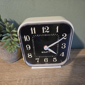 May include: A vintage, square-faced alarm clock with a black dial and white numerals. The clock has a white frame and the word "EQUITY" is printed on the face. The clock is set on a wooden surface.