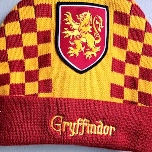 May include: A knitted beanie in red and yellow, featuring a checkered pattern and the word "Gryffindor" embroidered in gold. A patch with a gold lion on a red shield is sewn on the front.