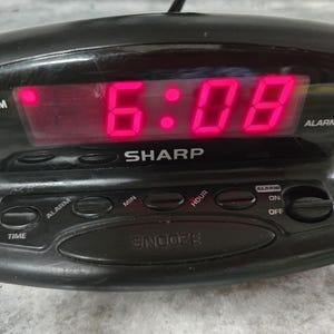 May include: Black Sharp digital alarm clock displaying 6:08 PM in bright red digits. Features buttons for alarm, time, minutes, and hours, with a snooze function. The clock has an alarm on/off switch.