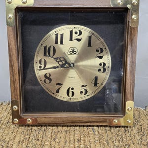 May include: Square wooden quartz clock with a gold-tone face and black background. The clock has a brown wooden frame with gold corner accents. The clock face features black numerals and hands. The words "QUARTZ" and "MADE IN USA" are visible.