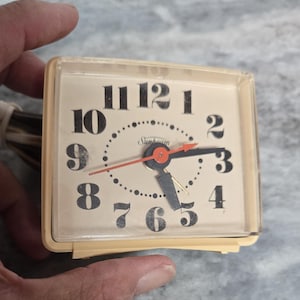 May include: A vintage Sunbeam alarm clock with a cream-colored face and a clear plastic frame. The clock face features large black numerals, black hands, and a red second hand. The clock is held in a hand.