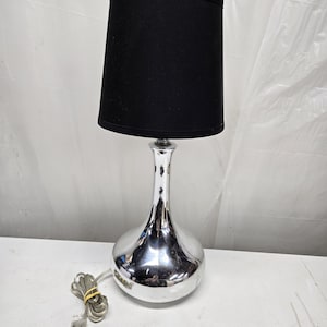 May include: A modern table lamp with a black lampshade and a shiny, chrome-colored base. The base has a bulbous bottom and a slender neck. The lamp is plugged in and ready to use. The lamp is a decorative lighting fixture.