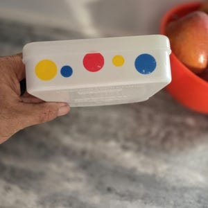 May include: A rectangular white plastic storage container featuring a series of colorful polka dots along the side. The pattern consists of alternating yellow, blue, and red circles of varying sizes printed on the smooth, opaque surface.