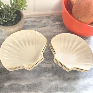 May include: A set of off-white, shell-shaped dishes. The dishes have a ribbed design and are stacked in sets of four. The background includes a small potted plant and a bowl of fruit.