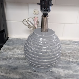 May include: A gray ceramic table lamp with a textured, scalloped design. The lamp has a silver metal base and a black socket.