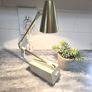 May include: A vintage desk lamp with a gold-colored conical shade and a cream-colored base. The lamp has an adjustable arm and a small potted succulent plant next to it. The lamp is plugged into a wall outlet.