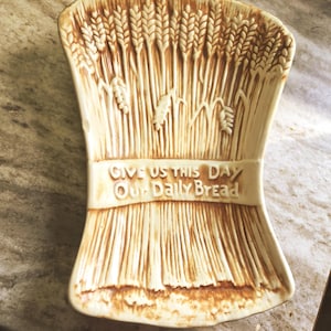 May include: A ceramic bread plate in the shape of a wheat sheaf, with a cream and brown color scheme. The plate features the embossed text "GIVE US THIS DAY OUR DAILY BREAD".