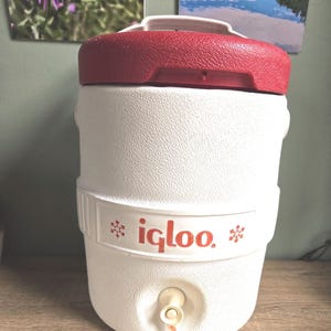 May include: A white Igloo water cooler with a red lid and handle. The cooler has a textured surface and a spigot at the bottom. The Igloo logo is printed in red on the side.