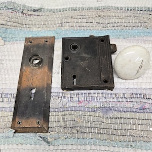 May include: An antique door hardware set featuring a rectangular backplate, a square lock mechanism, and a white oval doorknob. The metal components have a weathered, dark finish, showing signs of age and use. The set is likely from a vintage or historical home.