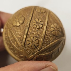 May include: A round, antique brass button with a detailed floral and geometric design. The button features embossed flowers and starburst patterns, with vertical lines and textured background. The button is held in a hand.