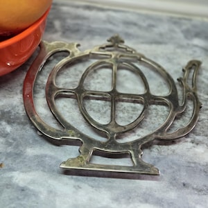 Puede incluir: Salvamanteles de plata con forma de tetera con un diseño intrincado. El salvamanteles tiene una forma circular con un centro elevado y un asa en el lateral.