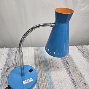 May include: A blue desk lamp with a flexible, chrome-colored neck and a round base. The lamp's shade is blue with an orange interior. The base has a switch and a sticker. The lamp is on a multi-colored rug.