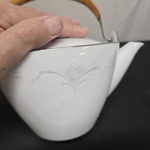May include: White porcelain teapot with a silver rim and a bamboo handle. The teapot features a floral embossed design on the side. The spout is curved, and the lid is silver. The teapot is on a black surface.