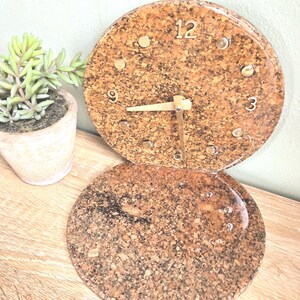 May include: A round cork clock with gold-colored hands and hour markers. The clock face features the numbers 12, 3, 6, and 9. A matching cork coaster sits below the clock. A small potted succulent plant is in the background.