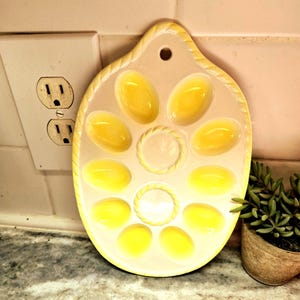 May include: A vintage ceramic egg holder with a pale yellow and cream color scheme. The oval-shaped tray has eight egg-shaped indentations and a central circular indentation. The edges are decorated with a rope-like design.