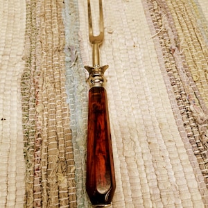 May include: A carving fork with a two-pronged stainless steel head and a dark reddish-brown wooden handle. The handle has a faceted design and is accented with a decorative metal collar. The fork is designed for serving meats.