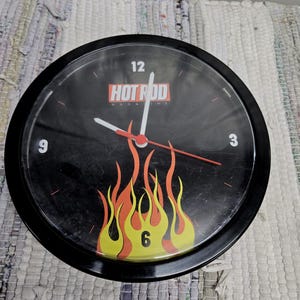 May include: Black round clock with the text "HOT ROD MAGAZINE" and a flame design in yellow, orange, and red. The clock has white hands and a red second hand. The numbers 3, 6, 9, and 12 are visible.