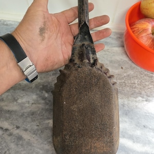 May include: A vintage, rusty metal garden trowel with a long handle and a rectangular blade. The trowel has a textured, aged appearance, suggesting it has been used for gardening. The handle is attached to the blade with a riveted design.