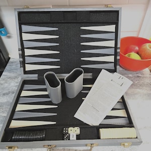 May include: Open backgammon set with a gray exterior and black interior. The game board features alternating white, gray, and black triangles. Includes dice, playing pieces, and dice cups. A sheet of instructions is also visible.