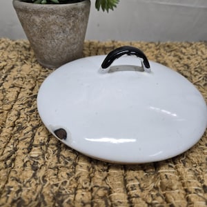 May include: A white, round lid with a black handle. The lid has a glossy finish and a small imperfection. A small potted plant is visible in the background.