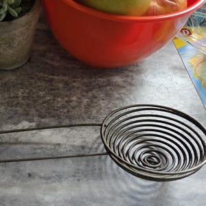 May include: A metal spiral skimmer with a long handle rests on a gray countertop. The skimmer's circular head features concentric wire coils. A red bowl with fruit and a potted plant are in the background.