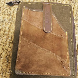 May include: A brown and green wallet with a suede pocket and a leather strap. The wallet has yellow stitching and a zipper closure. The leather strap has the word "VAAN" printed on it.