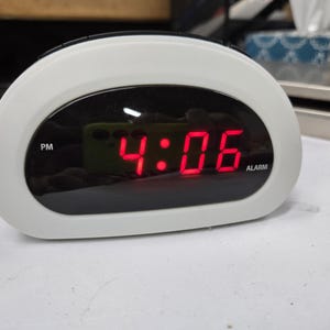 May include: White and black digital alarm clock displaying 4:06 PM in red numerals. The clock has a curved design with an oval-shaped display. The word "ALARM" is visible on the display.