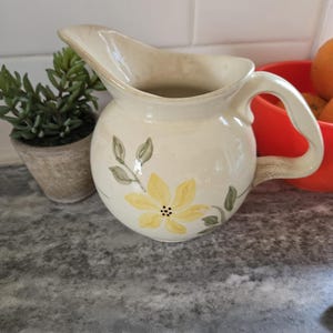 May include: A white ceramic pitcher with a yellow flower design. The pitcher has a handle and a spout. The pitcher is sitting on a gray countertop.