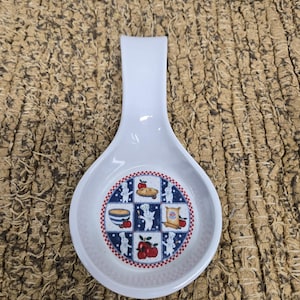 May include: White ceramic spoon rest with a wide, oval bowl and a handle. The bowl features a circular design with illustrations of food items and a chef. The design is framed by a red and blue border.