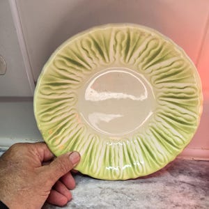 May include: A ceramic plate with a cabbage leaf design. The plate has a light green and cream color scheme, with a scalloped edge. The plate is round and has a textured surface, with the cabbage leaf pattern radiating from the center.