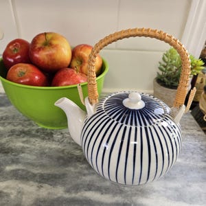 May include: A white teapot with a blue striped pattern and a woven bamboo handle. The teapot is sitting on a gray countertop.