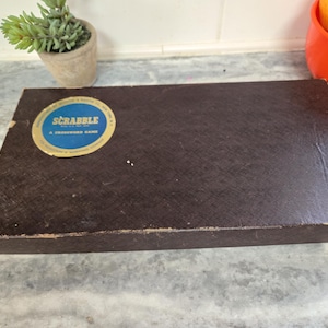 May include: A brown Scrabble board game box with a blue and gold circular label. The box is rectangular and sits on a gray surface. A small potted plant and a red bowl with fruit are in the background.