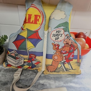 May include: Vintage ALF life vest with a yellow interior and a light blue exterior featuring beach scenes and the ALF logo. The vest has a zipper closure and adjustable straps. The graphic shows ALF on a beach chair with the text "Yo! Surf's Up".