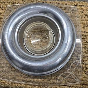 Puede incluir: Una lámpara de metal plateado con un centro de vidrio transparente, colocada sobre una base cuadrada transparente con diseños en forma de hoja. La lámpara tiene un diseño circular con anillos concéntricos.