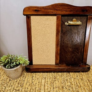 May include: A dark stained wooden memo board with a corkboard section and a dark wood section with a gold-colored clip. The memo board has a decorative top and is sitting on a woven surface.