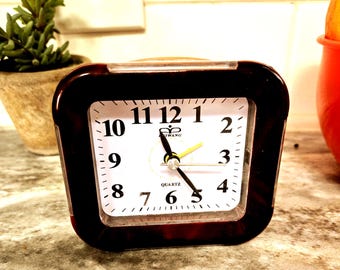 May include: A brown and black quartz analog alarm clock with a white face. The clock has black numbers and hands, with a yellow second hand. The clock is on a countertop.
