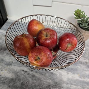 May include: A silver wire fruit bowl filled with five red apples. The bowl is on a gray countertop.