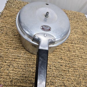 May include: A vintage silver-colored Presto pressure cooker with a black handle. The cooker has a round shape with a lid and a pressure release valve. The Presto logo is visible on the front.