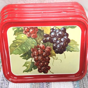 May include: A stack of vintage red metal trays. Each tray features a rectangular design with rounded corners and a painted image of grapes and leaves in shades of red, purple, and green on a cream background.