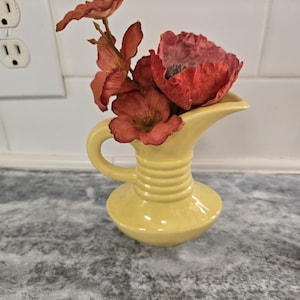May include: A yellow ceramic pitcher with a wide mouth and a handle, filled with a bouquet of red and orange artificial flowers.
