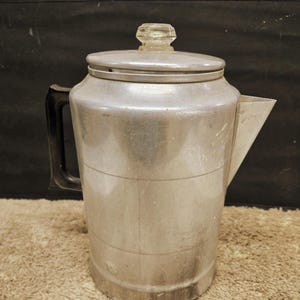 May include: A vintage aluminum percolator coffee pot featuring a cylindrical body with horizontal etched lines, a black plastic handle, a triangular spout, and a removable lid topped with a clear, faceted glass knob for viewing the coffee.