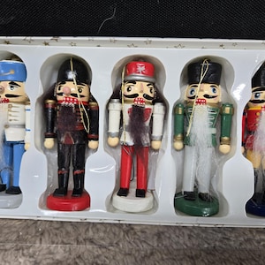 May include: A set of five small nutcracker ornaments in a white plastic tray. Each figure features a painted uniform, a black or red hat, and a white faux fur beard. The nutcrackers are colored in blue, black, red, green, and yellow with gold hanging cords.