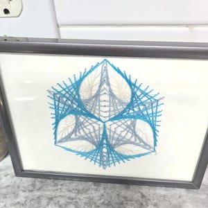 May include: Framed geometric string art in a gray frame. The artwork features a symmetrical design with blue and gray threads on a white background. The frame is rectangular and has a dark gray finish. The artwork is displayed on a light-colored surface.