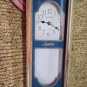 May include: A rectangular wooden clock with a blue face and a gold-colored trim. The clock face displays the Ingraham brand name and the time. Below the clock is a space for a photo. The clock is marked "USA".