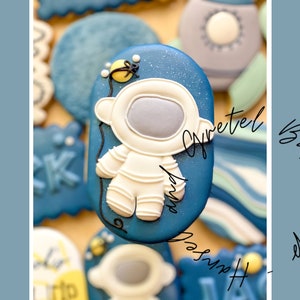 Può includere: Un biscotto a forma di astronauta blu e bianco decorato con una stella gialla. Il biscotto ha una forma ovale e un contorno bianco.
