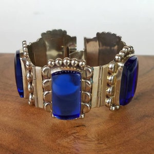 May include: A silver-toned bracelet with rectangular, deep blue glass stones. The bracelet has a hinged design with decorative metalwork and is displayed on a wooden surface. The bracelet's design features a series of rectangular blue stones.