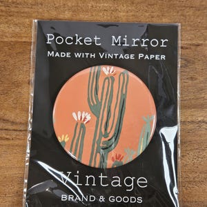 May include: A round pocket mirror featuring a terracotta orange background with a green cactus illustration and white flower accents. The mirror is packaged in a clear plastic sleeve with a black card labeled Pocket Mirror Made with Vintage Paper.